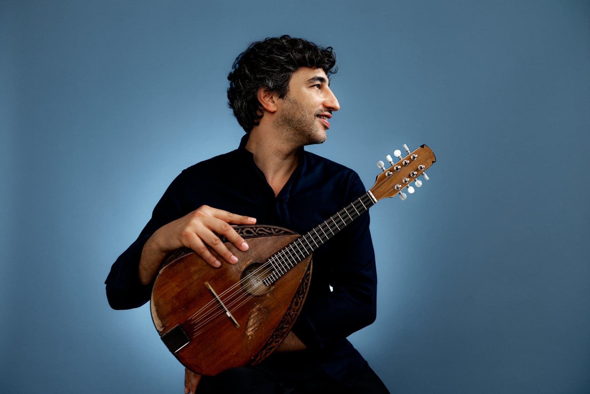 Avi Avital heldur á mandólíni og horfir til hliðar, á bláum bakgrunni.