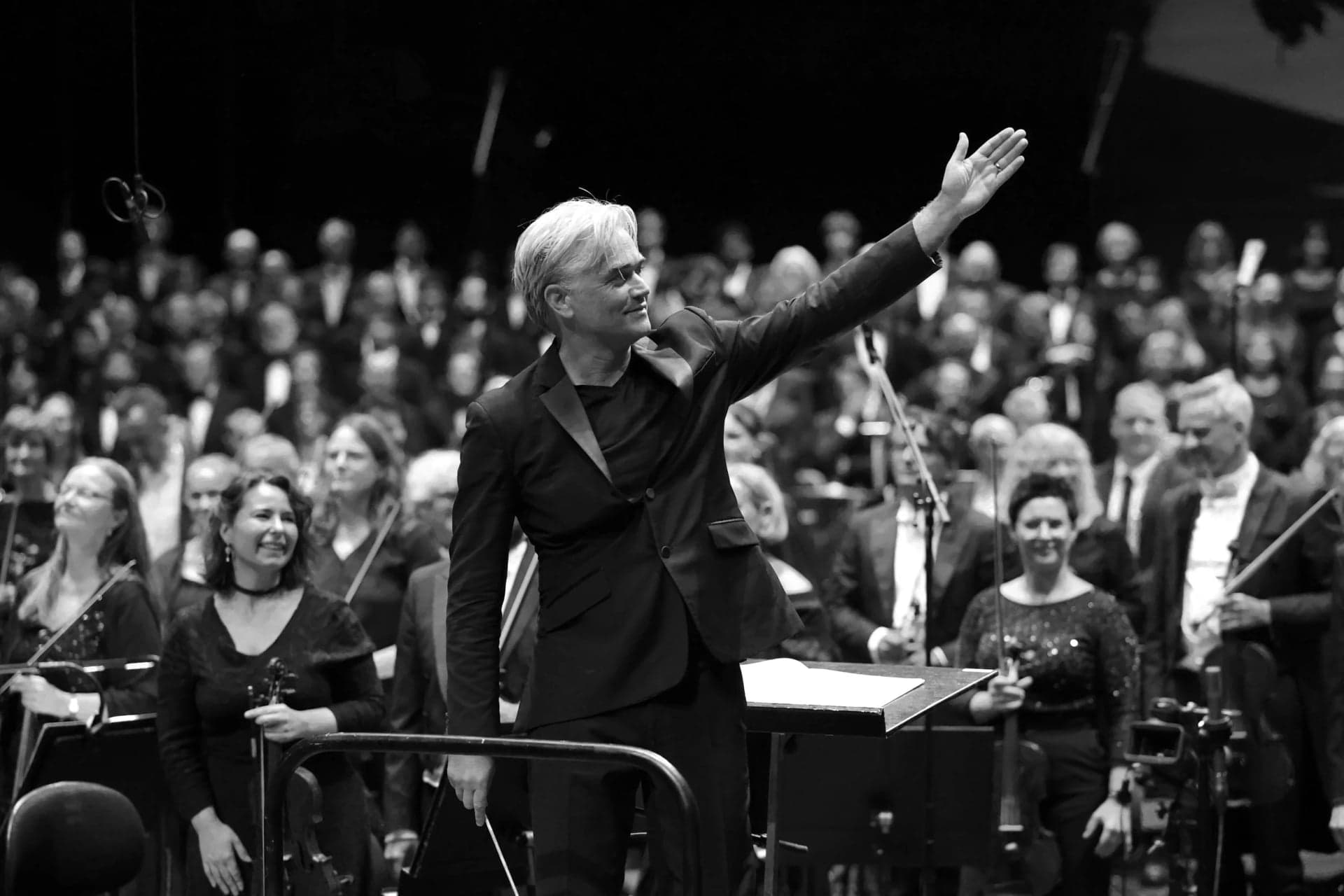 Edward Gardner raises an arm towards the performers behind him, in black and white.