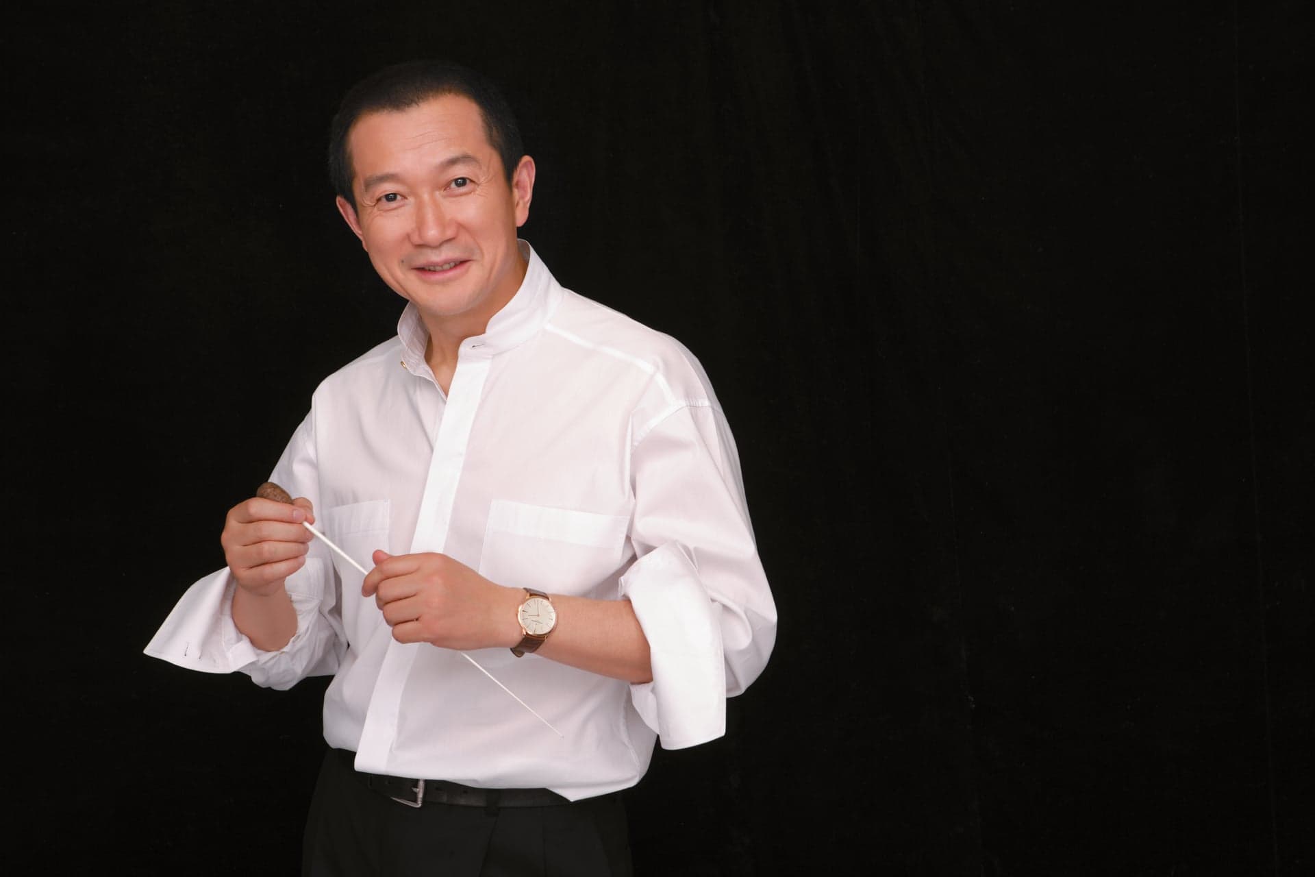 Tan Dun holds a baton in both hands, in a white shirt against a black background.
