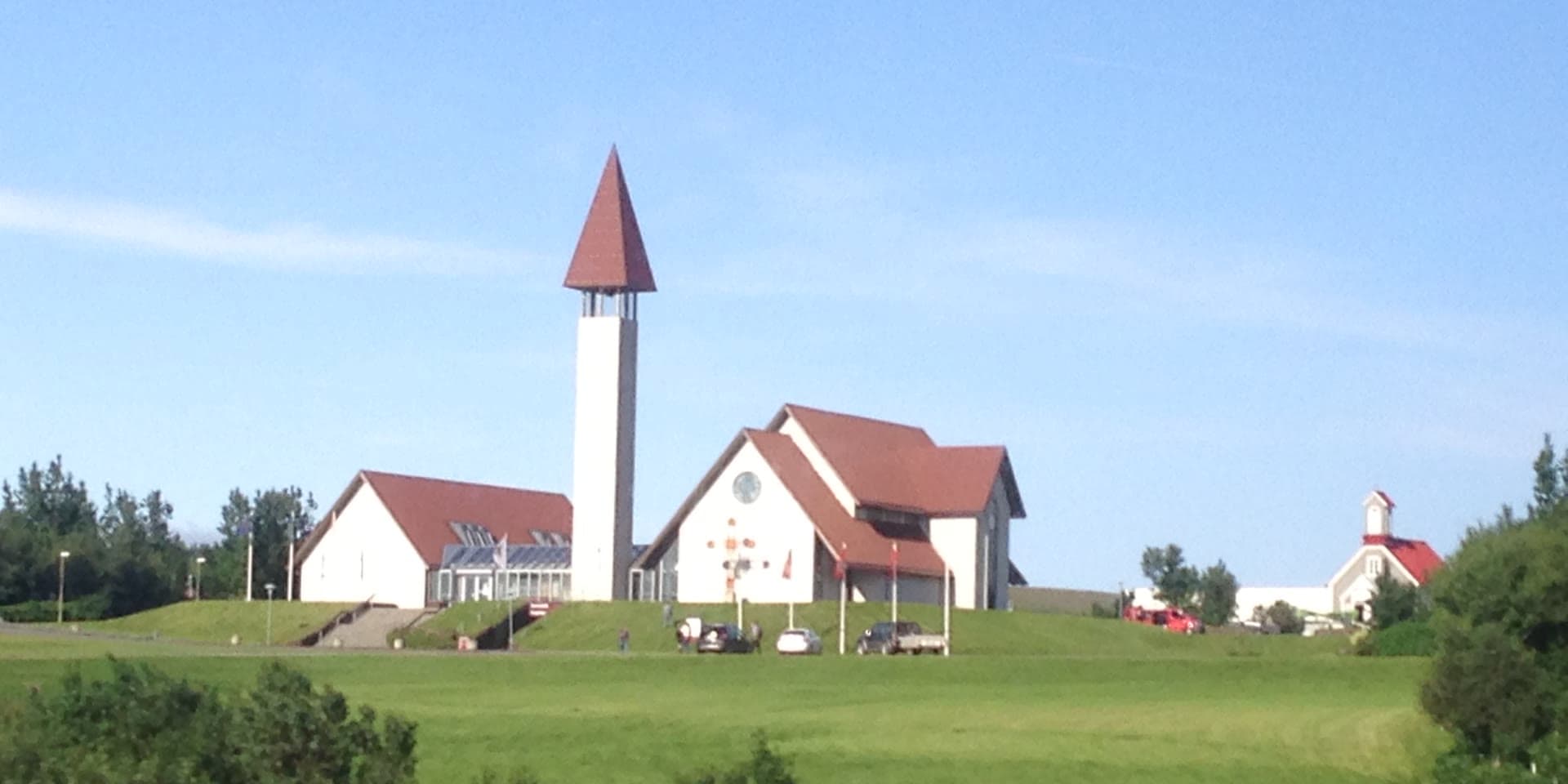Reykholtskirkja á hæð sinni í Reykholti, hvít með rauðum þökum og háum, mjóum klukkuturni, og gamla timburkirkjan til hægri innan um trén.