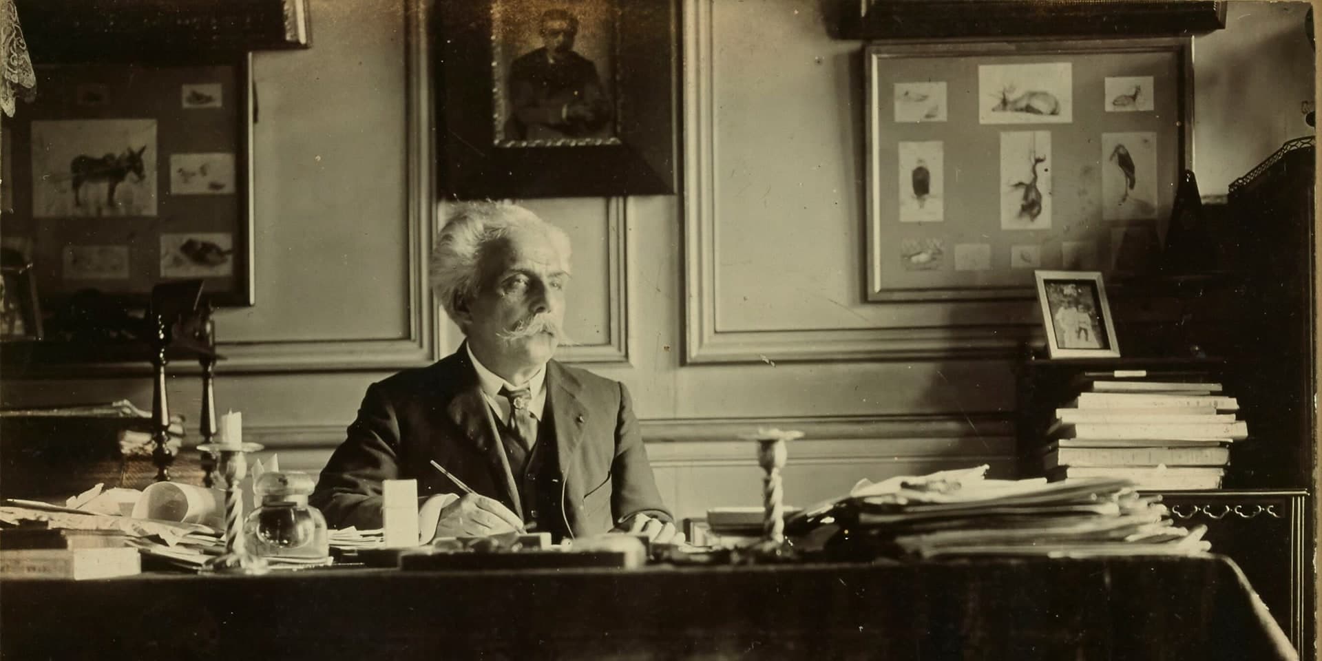 Gabriel Fauré at his desk with a pen in his hand, in a room hung with framed drawings.
