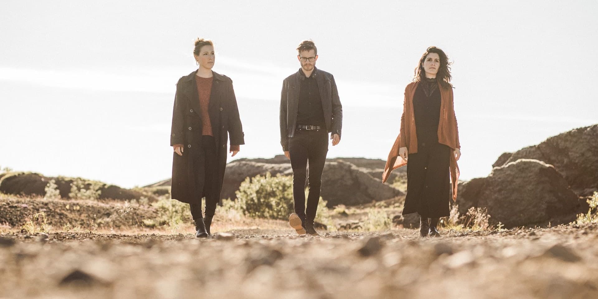 The three members of KIMI ensemble walking side by side along a gravel track through rocky ground under a bright sky.