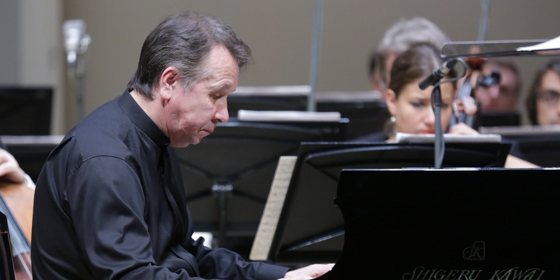 Mikhail Pletnev in a black shirt at a grand piano, with players of an orchestra behind him.