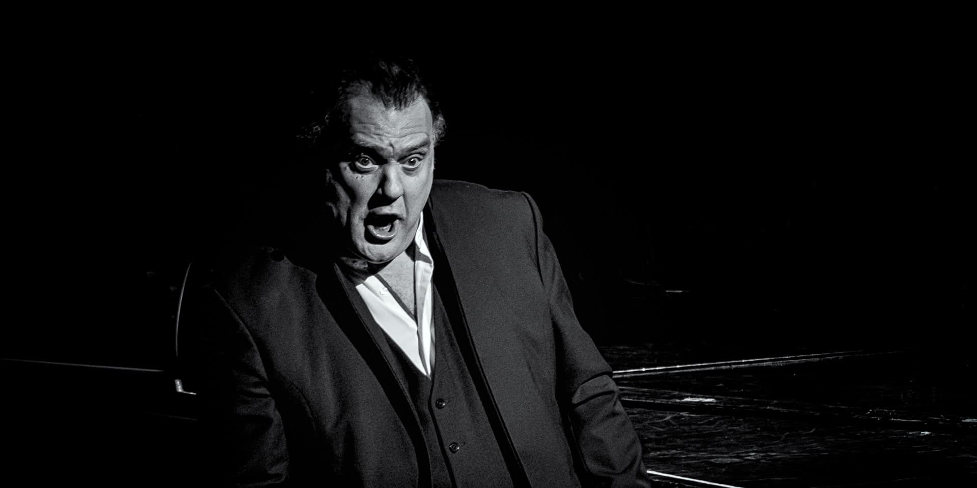 Bryn Terfel singing on a dark stage beside a grand piano.