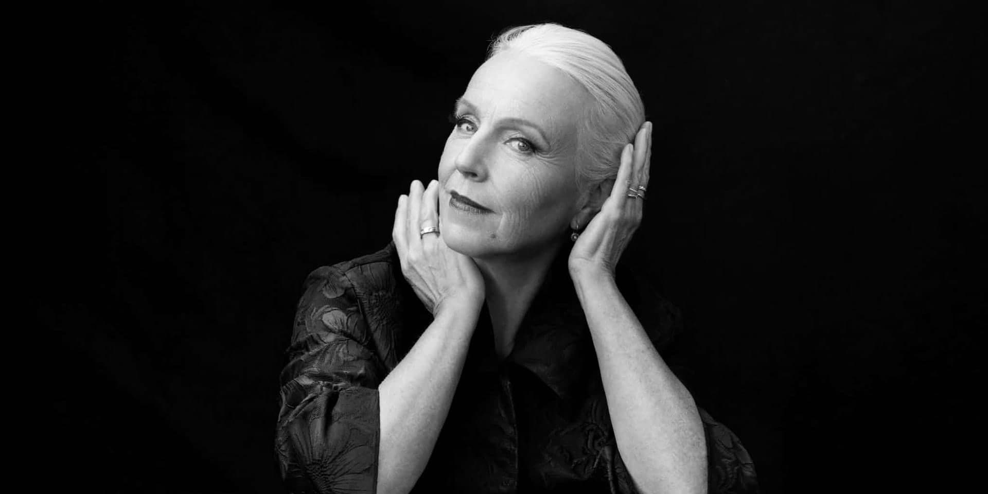 Anne Sofie von Otter seated at a table against a dark ground, her head resting in her hands.