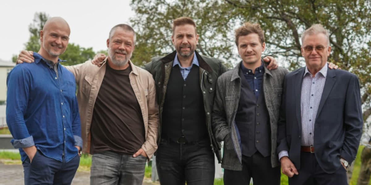 Five men standing together outdoors with their arms round each other's shoulders, trees behind them.