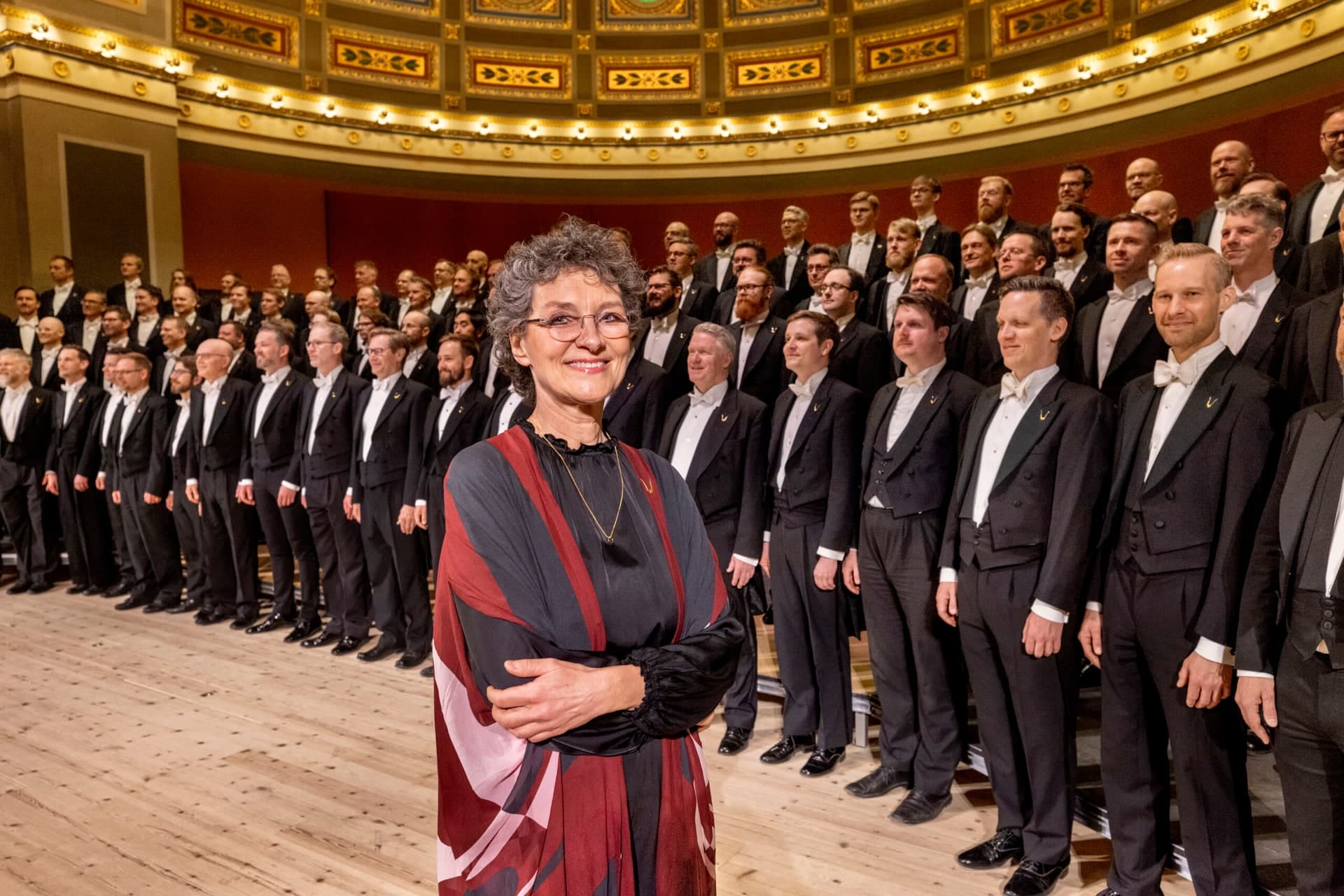 The choir stands in ranks on a stage in white tie, with Cecilia Rydinger at the front, arms folded, in a dark robe with red panels.
