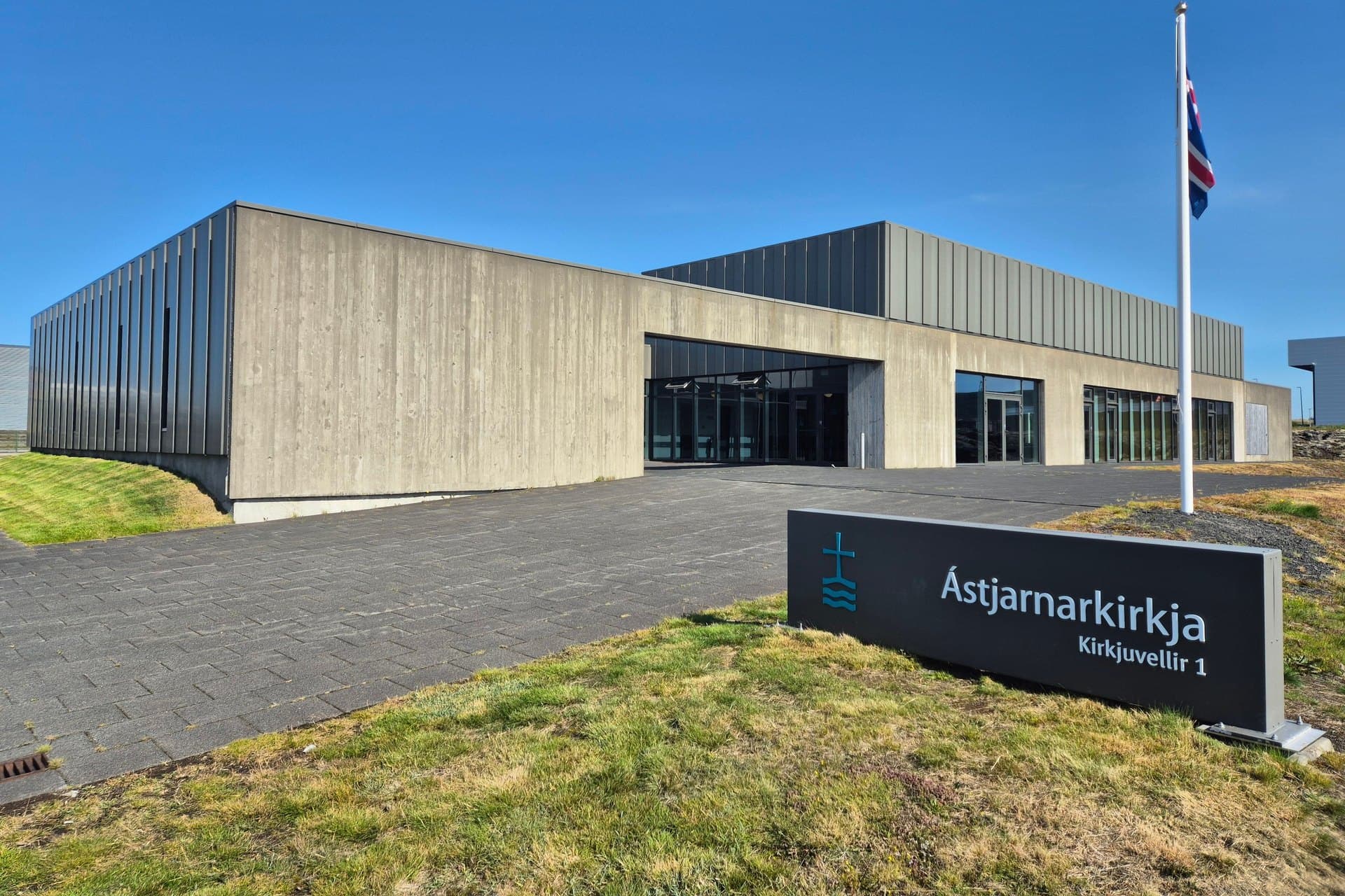 Ástjarnarkirkja, lágt steinsteypt hús með glerjuðum inngangi og skilti með nafni kirkjunnar á grasinu fyrir framan.