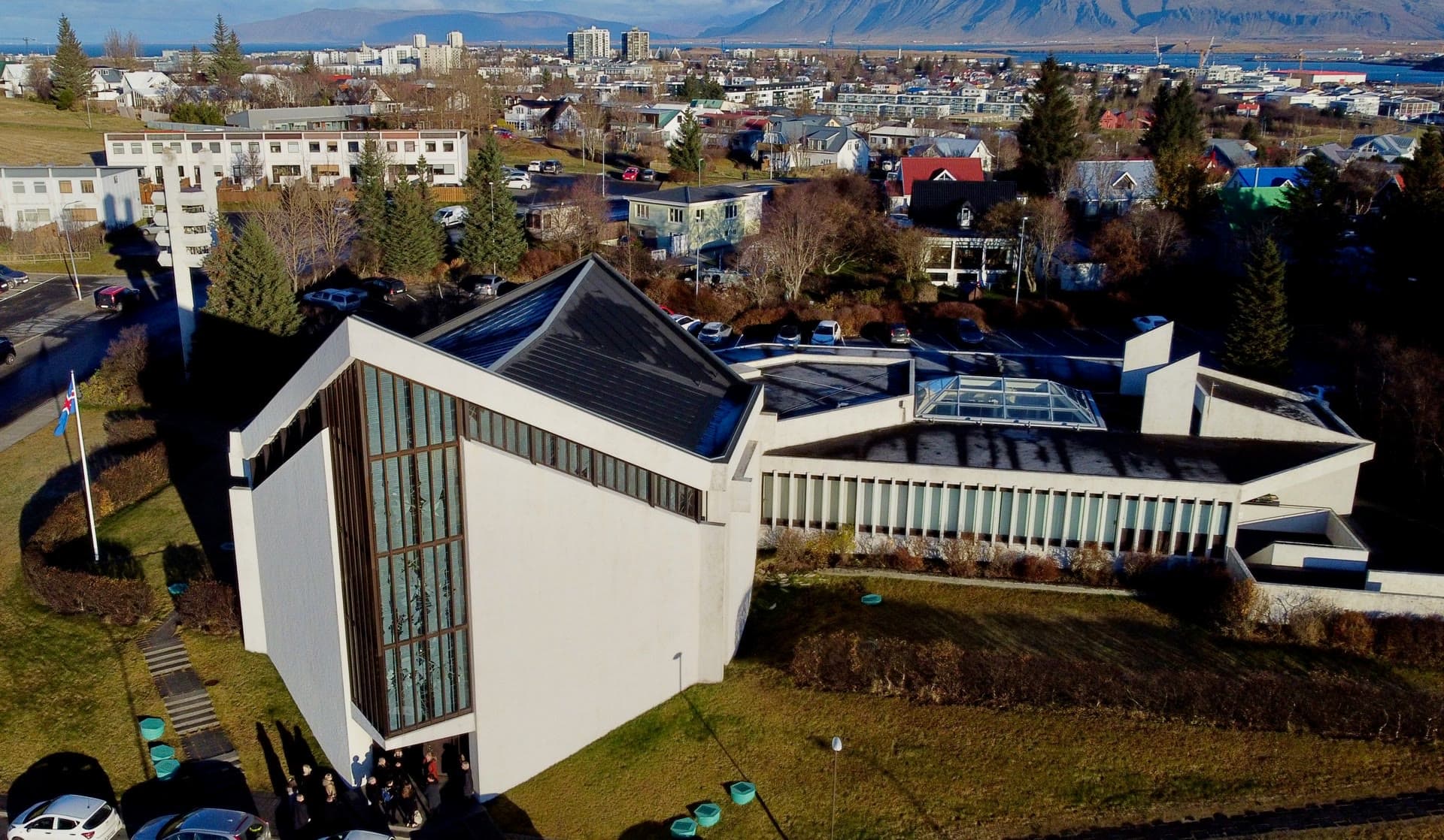 Bústaðakirkja from the air, the pitched roof of the nave and the long parish hall beside it, with the city and Esja behind.
