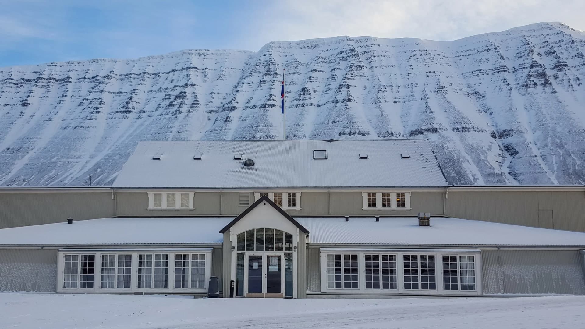 Framhlið Edinborgarhússins í snjó, gaflbyggður inngangur á miðju með glerjuðum boga yfir dyrunum, raðir hvítra smárúðuglugga til beggja handa, íslenski fáninn á stöng upp af mæninum og snævi þakin fjallshlíð að baki.