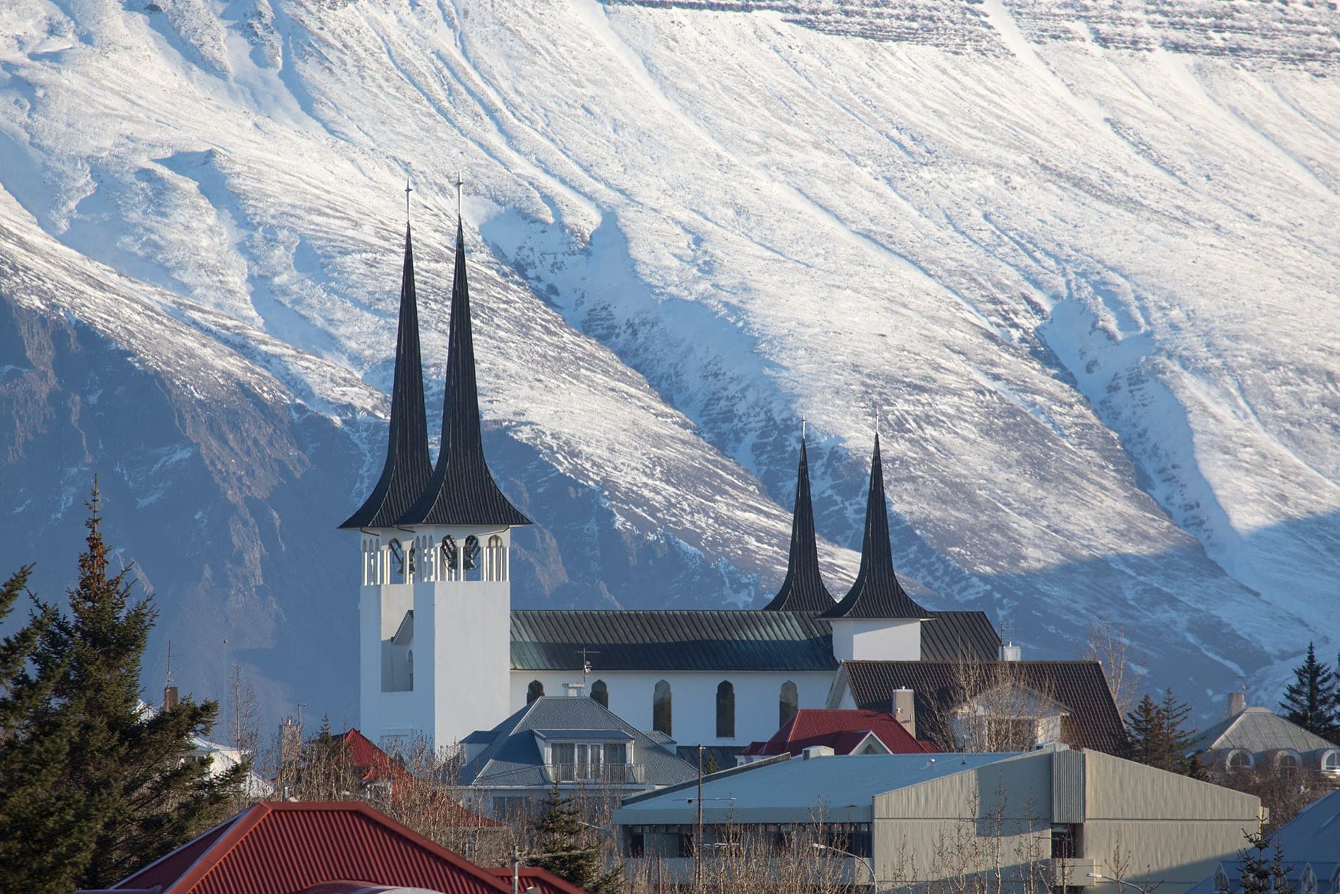 Háteigskirkja með fjórum svörtum turnspírum yfir hvítri framhlið, snævi þakin fjallshlíð fyllir bakgrunninn og húsþök standa fyrir framan.