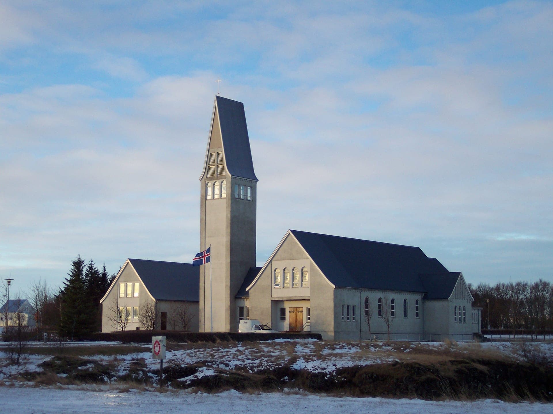 Selfosskirkja í vetrarbirtu, hár turn með krossi og bröttum þökum til beggja handa, íslenskur fáni á stöng fyrir framan og snjór í grasinu.