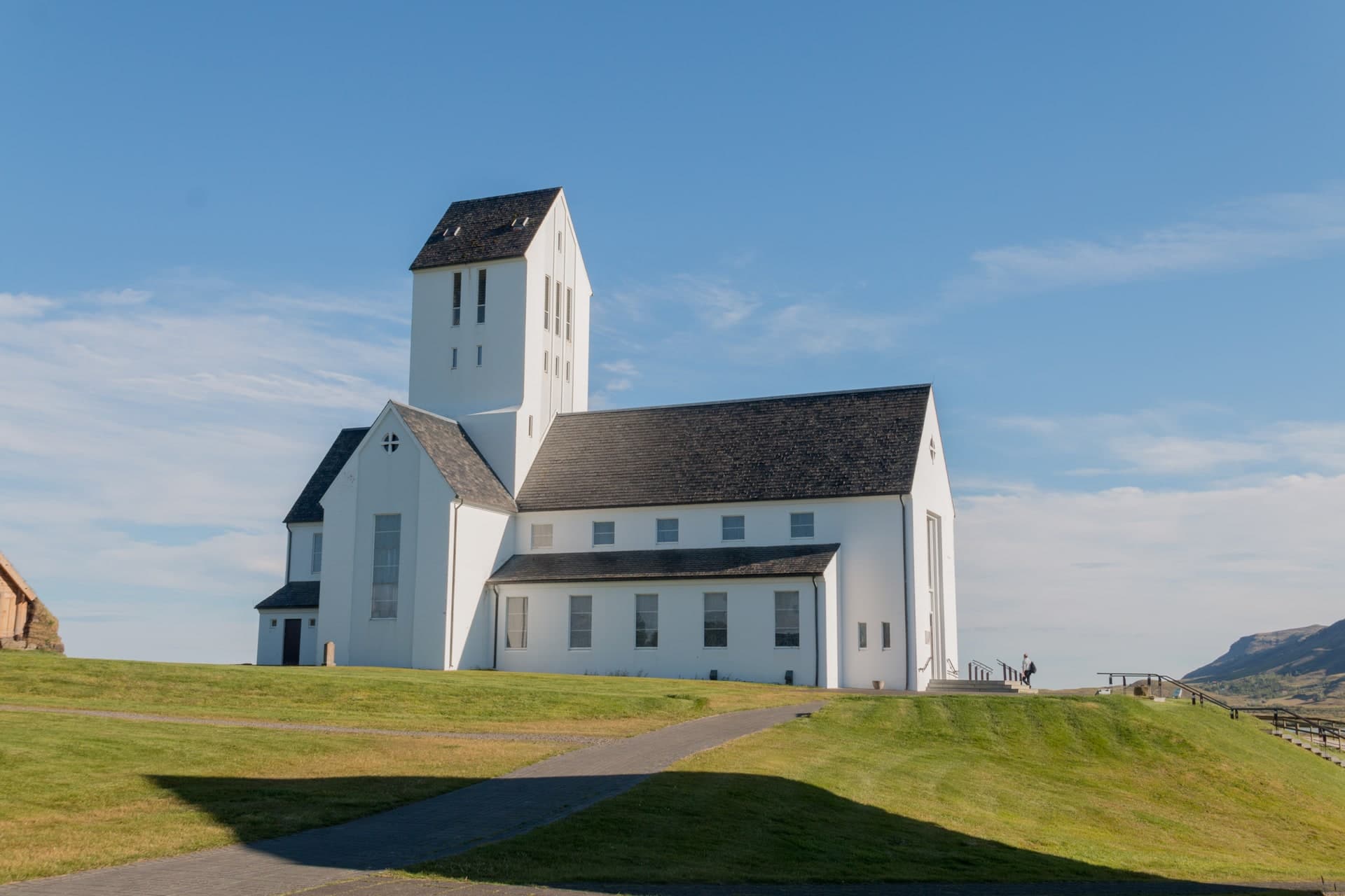 Skálholtsdómkirkja in sunshine, white with a dark roof and a tall tower, a path running up the grass slope to the door.