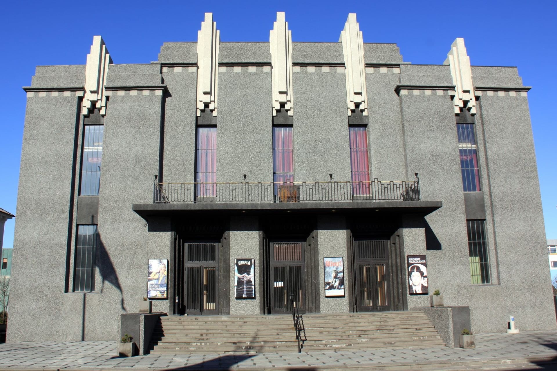 The front of the National Theatre on Hverfisgata in sunshine, a grey rendered building with pale stepped piers rising above the roofline, a balcony over three doorways and broad steps down to the pavement.