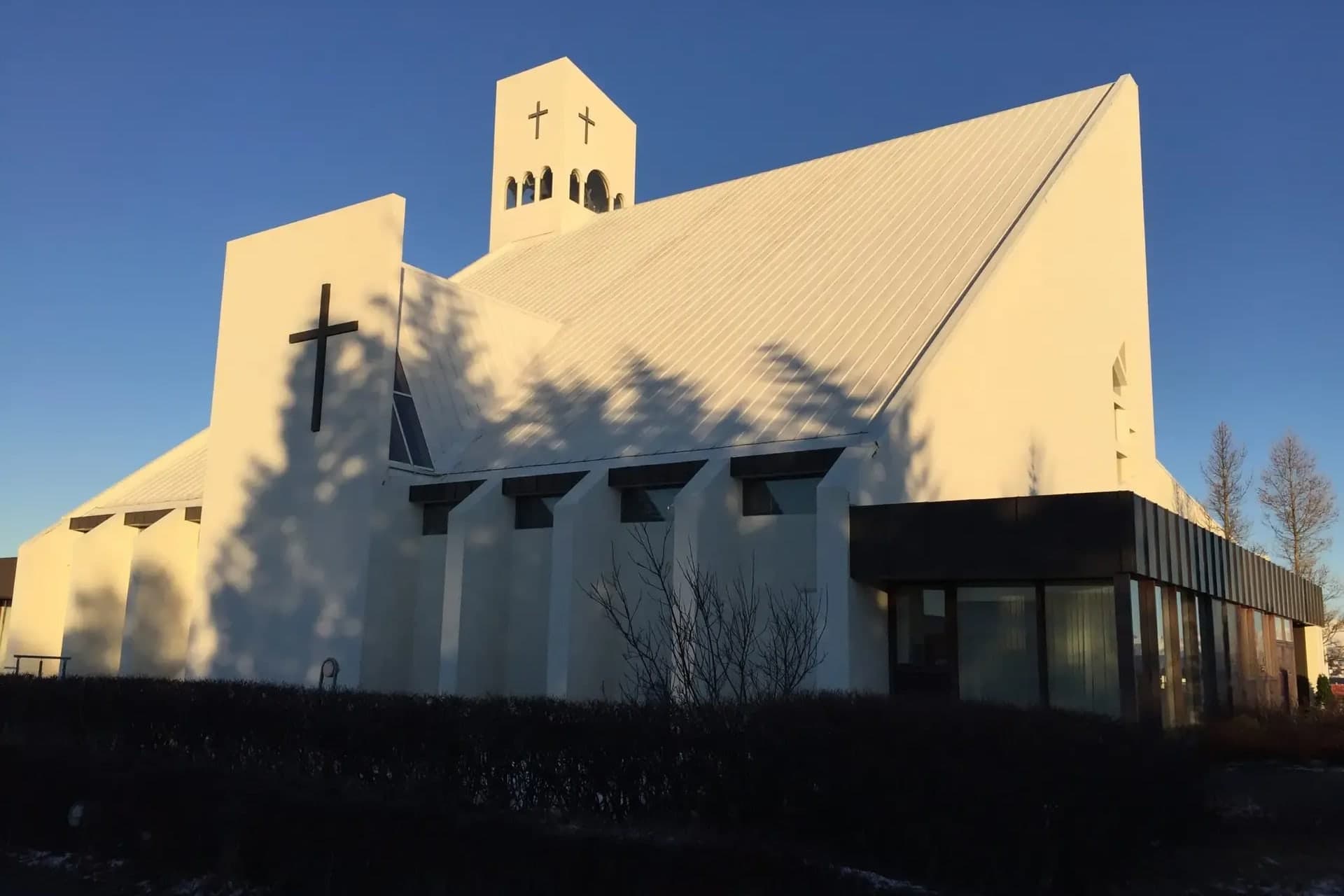 Vídalínskirkja í lágri vetrarsól, hvít kirkja með bröttu þaki, hvítum klukknaturni með tveimur krossum og stórum dökkum krossi á hvítum vegg.