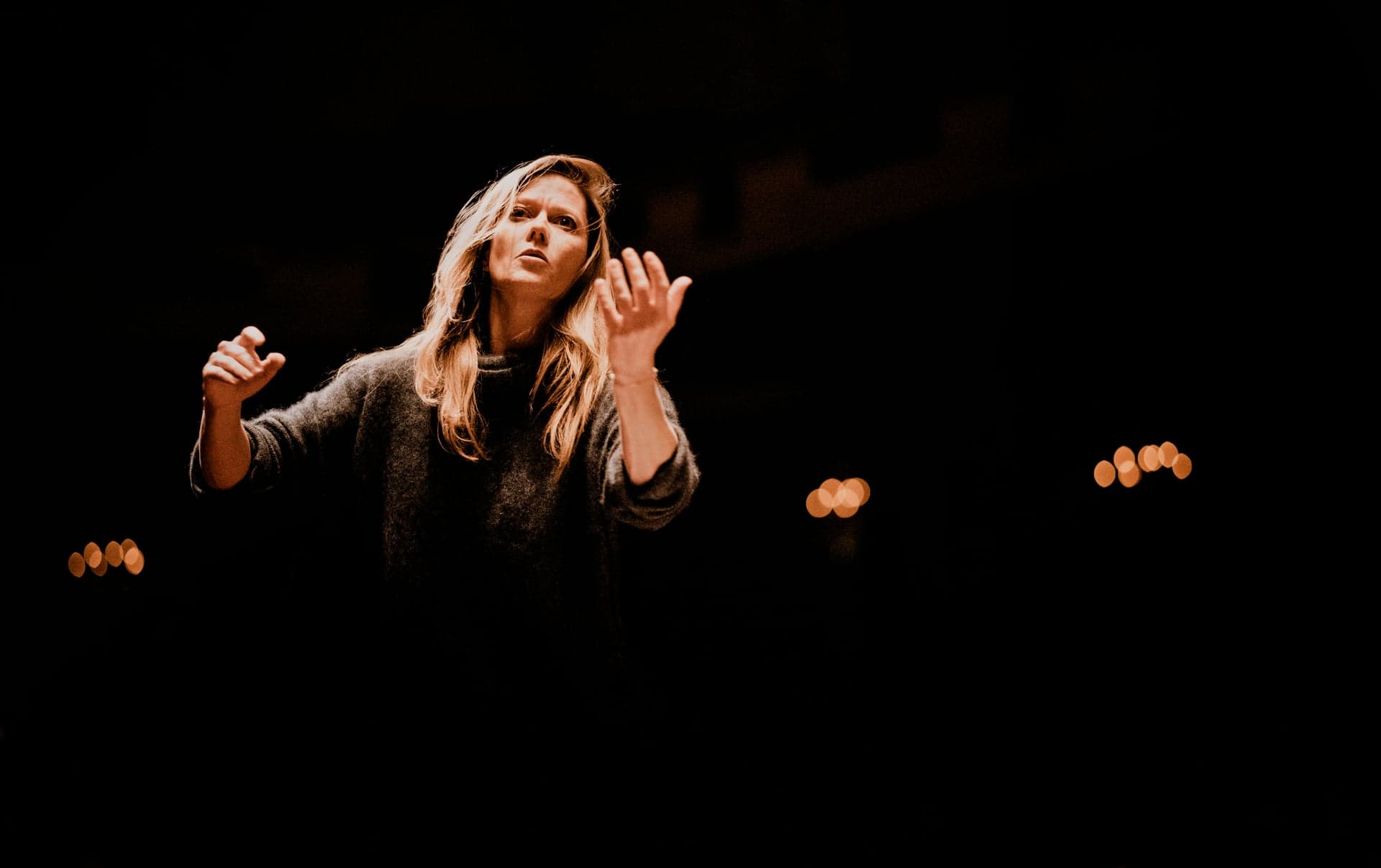 Barbara Hannigan conducts on a dark stage, lit from the front with both hands raised.