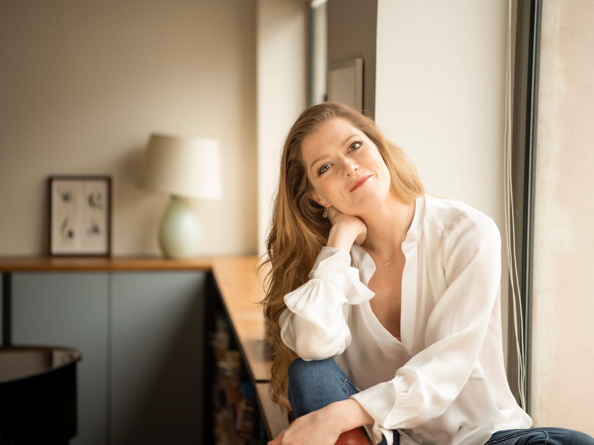 Barbara Hannigan sits by a window in a white shirt with her chin resting in her hand.