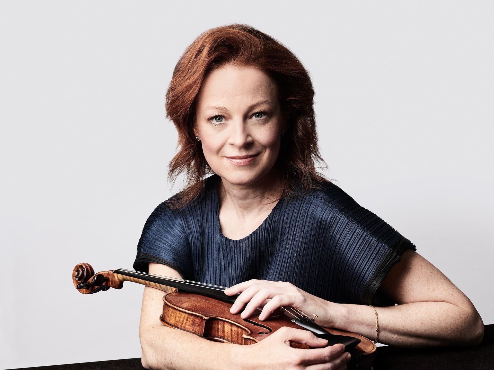 Carolin Widmann holds a violin and smiles, against a grey background.