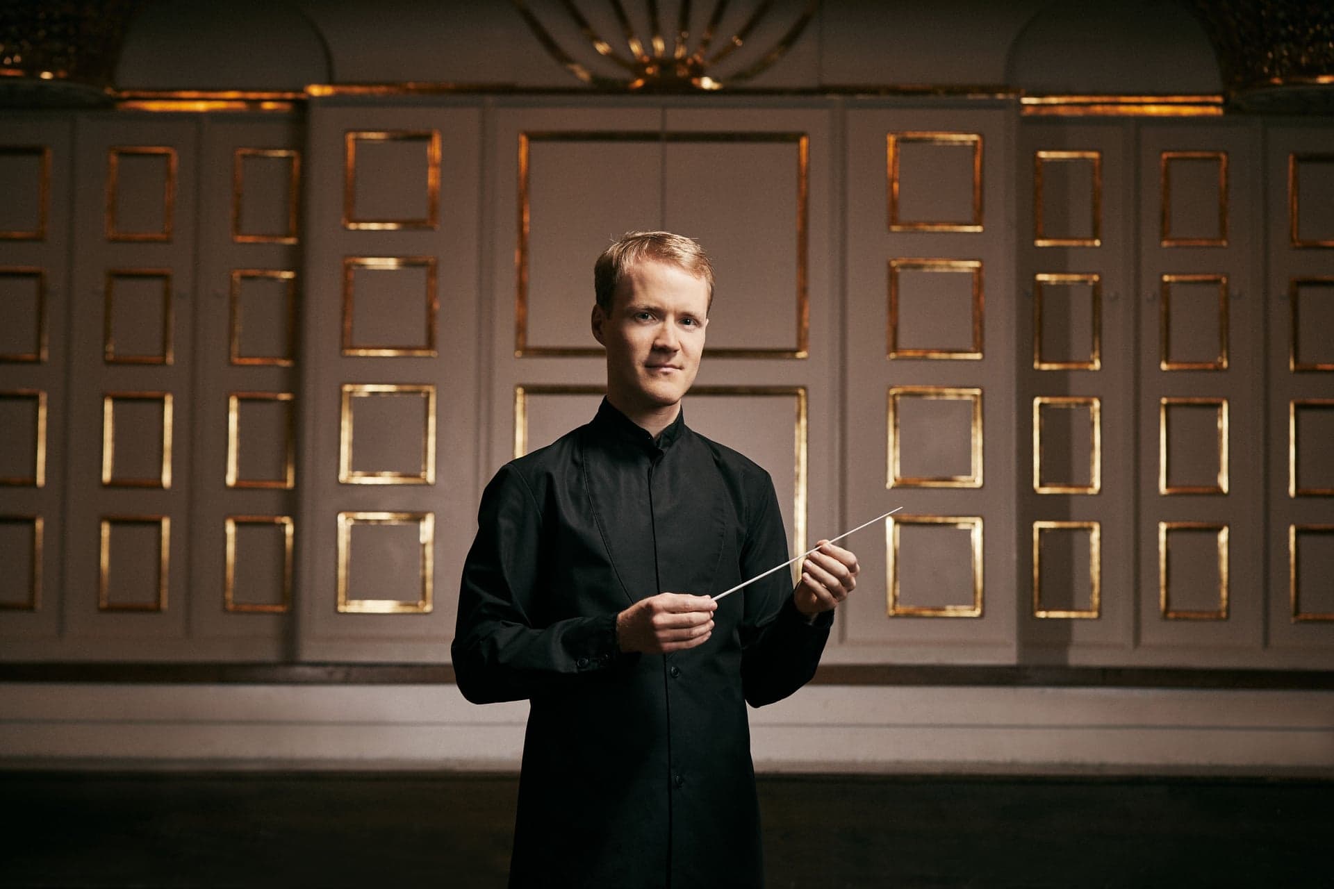 Christoph Koncz stands holding a baton in front of gilded wall panels.