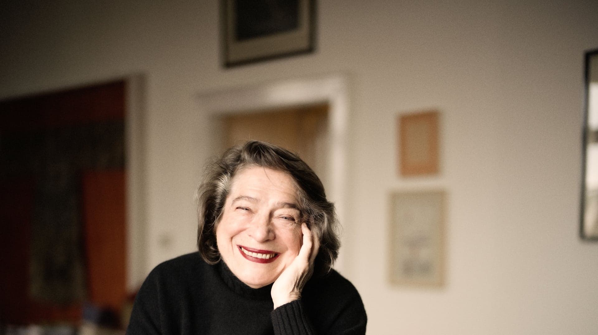 Elisabeth Leonskaja laughs with a hand at her cheek, in a room hung with pictures.
