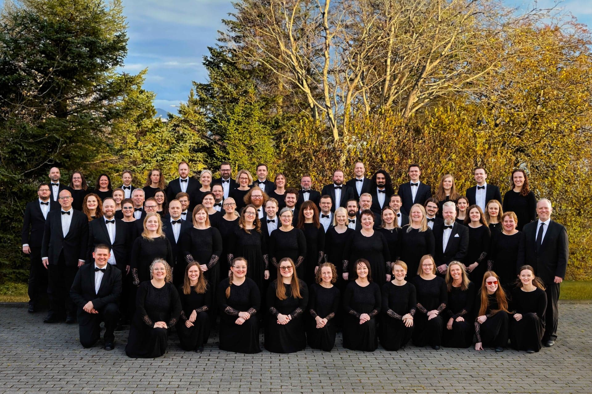 Söngsveitin Fílharmónía in black, standing outdoors in rows in front of trees.