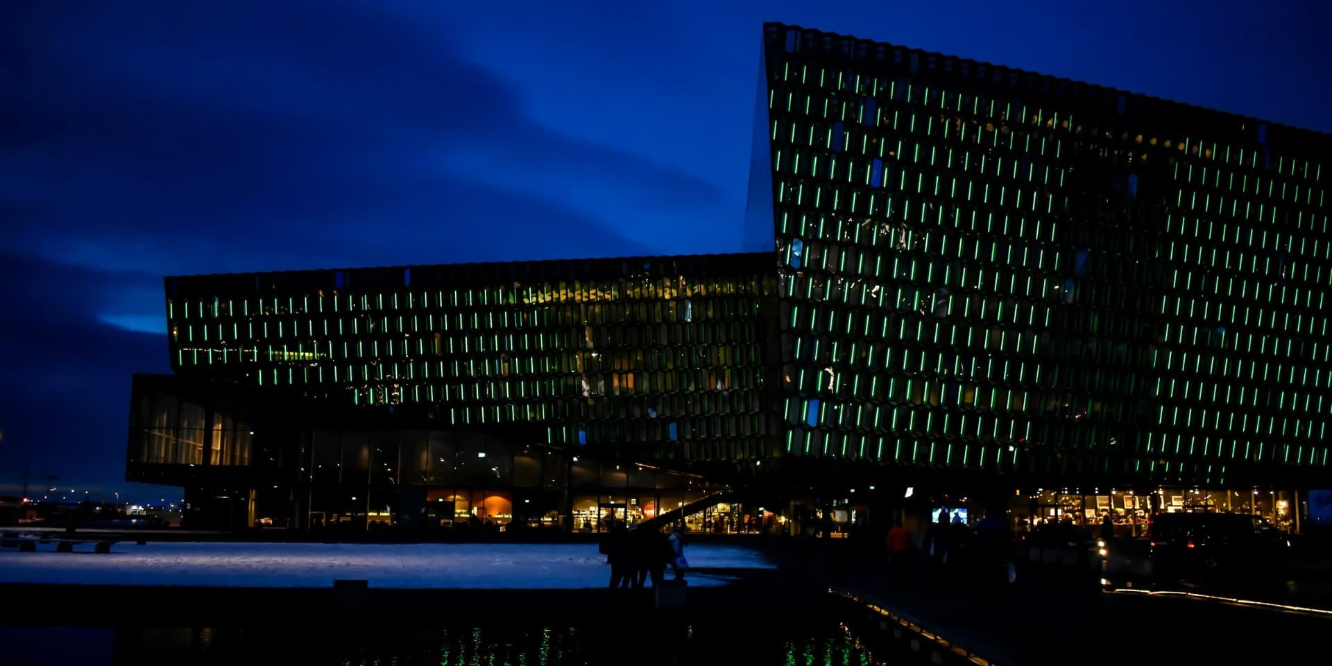 Tónlistarhúsið Harpa í Reykjavík á bláa tímanum á vetrarkvöldi, glerhjúpurinn glóir í grænum ljósum yfir snjó og dimmu vatni.