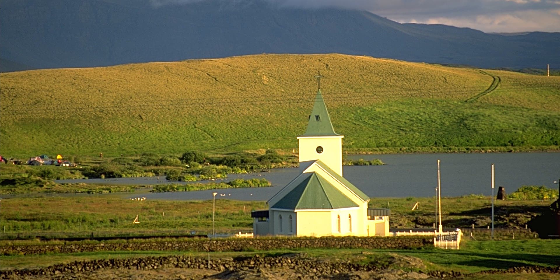 Reykjahlíðarkirkja in evening light, white with a green roof and tower, the lake and a grassy hillside behind it.