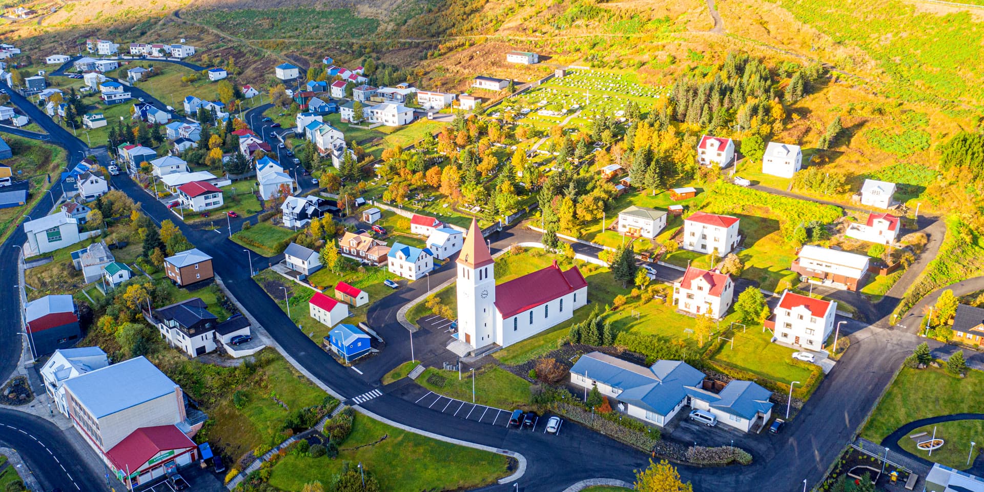 Siglufjörður from the air in autumn, Siglufjarðarkirkja white with a red roof at the centre, among houses and yellowing trees.