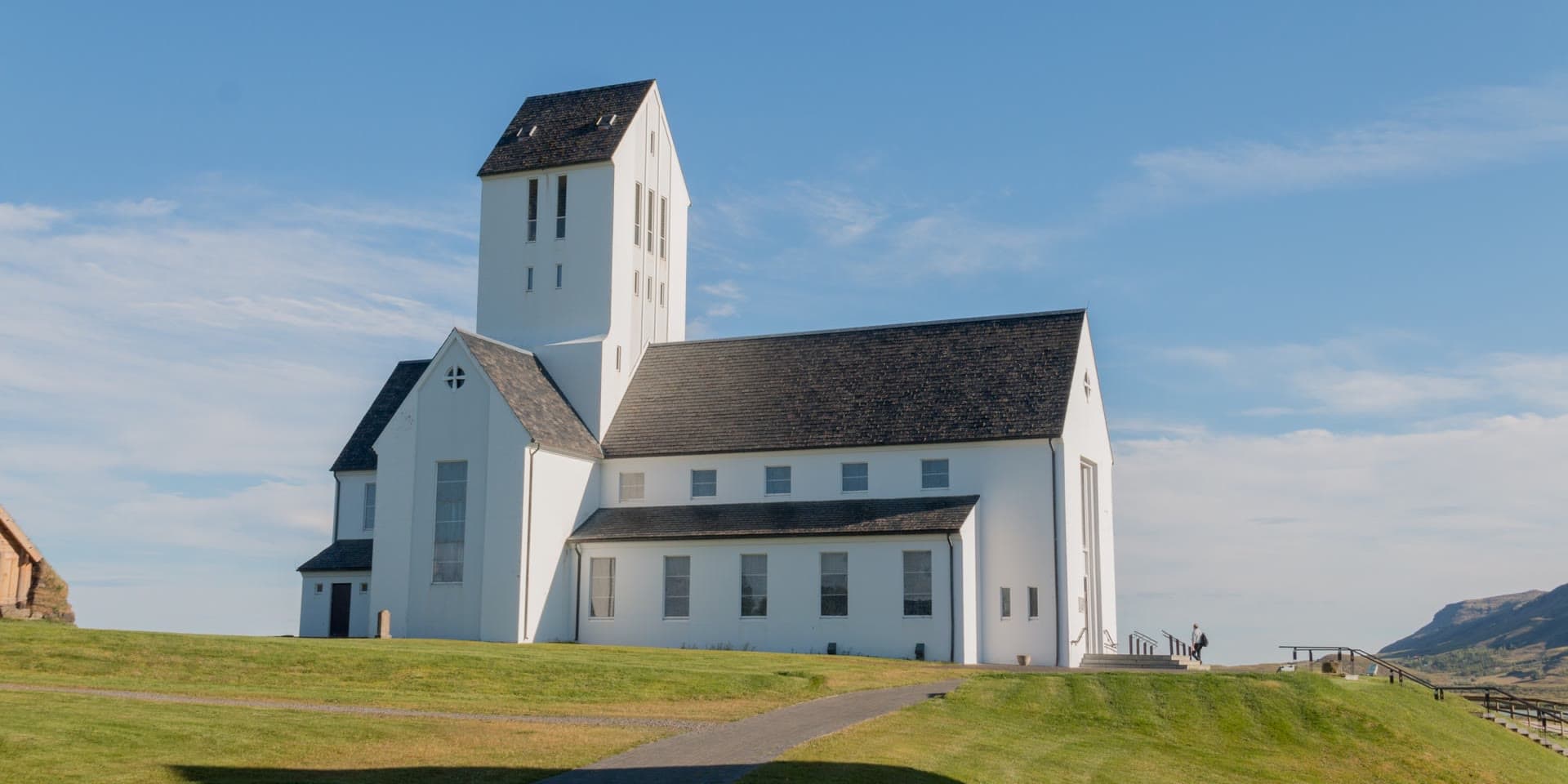Skálholtsdómkirkja in sunshine, white with a dark roof and a tall tower, a path running up the grass slope to the door.