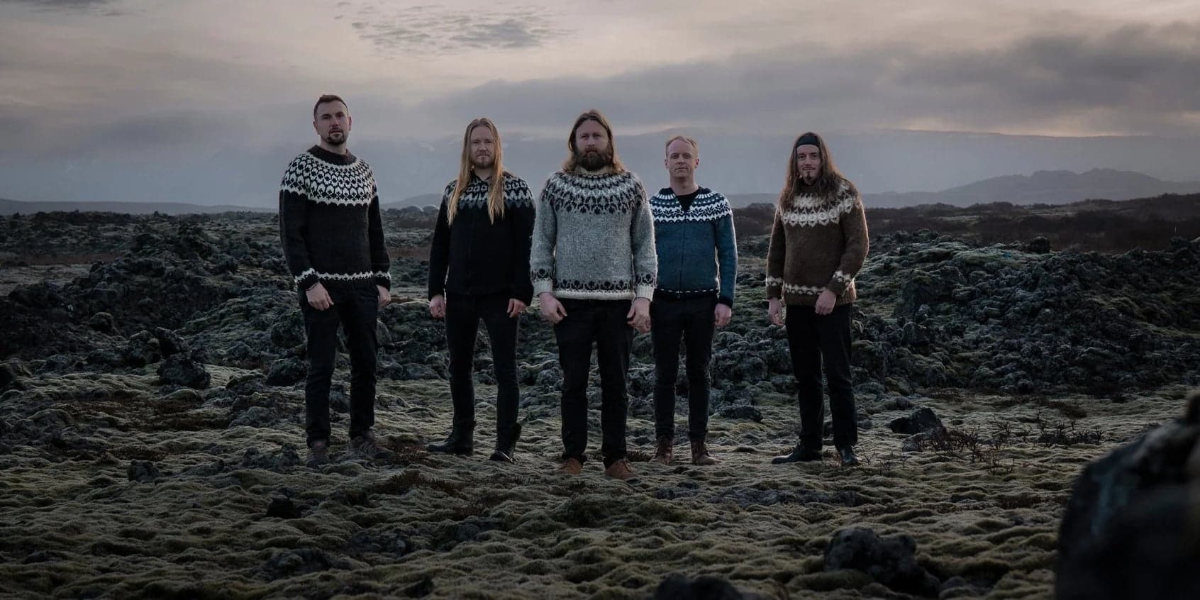 Five men in Icelandic sweaters standing in a mossy lava field, with mountains and a clouded sky behind them.