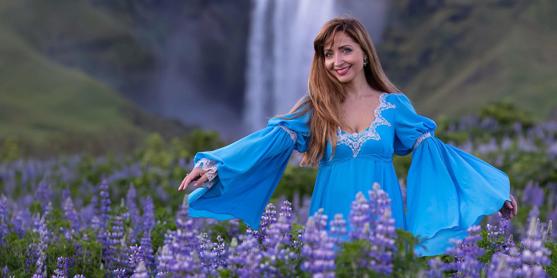 Alexandra Chernyshova standing in a field of lupins with her arms spread, smiling, a waterfall behind her.