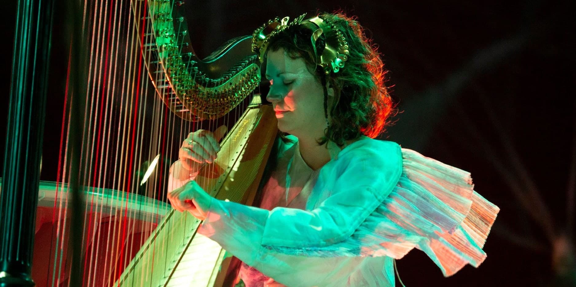 A harpist plays a concert harp under coloured stage light, her eyes closed and a gilded ornament in her hair.