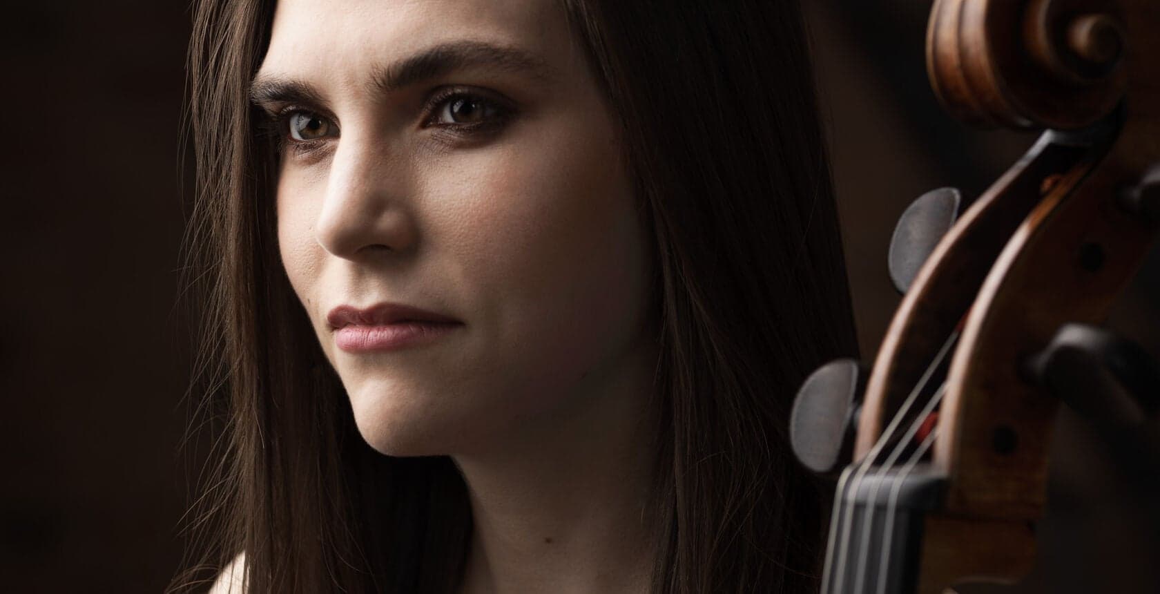 A young woman looks at the camera with the scroll and strings of a cello beside her.