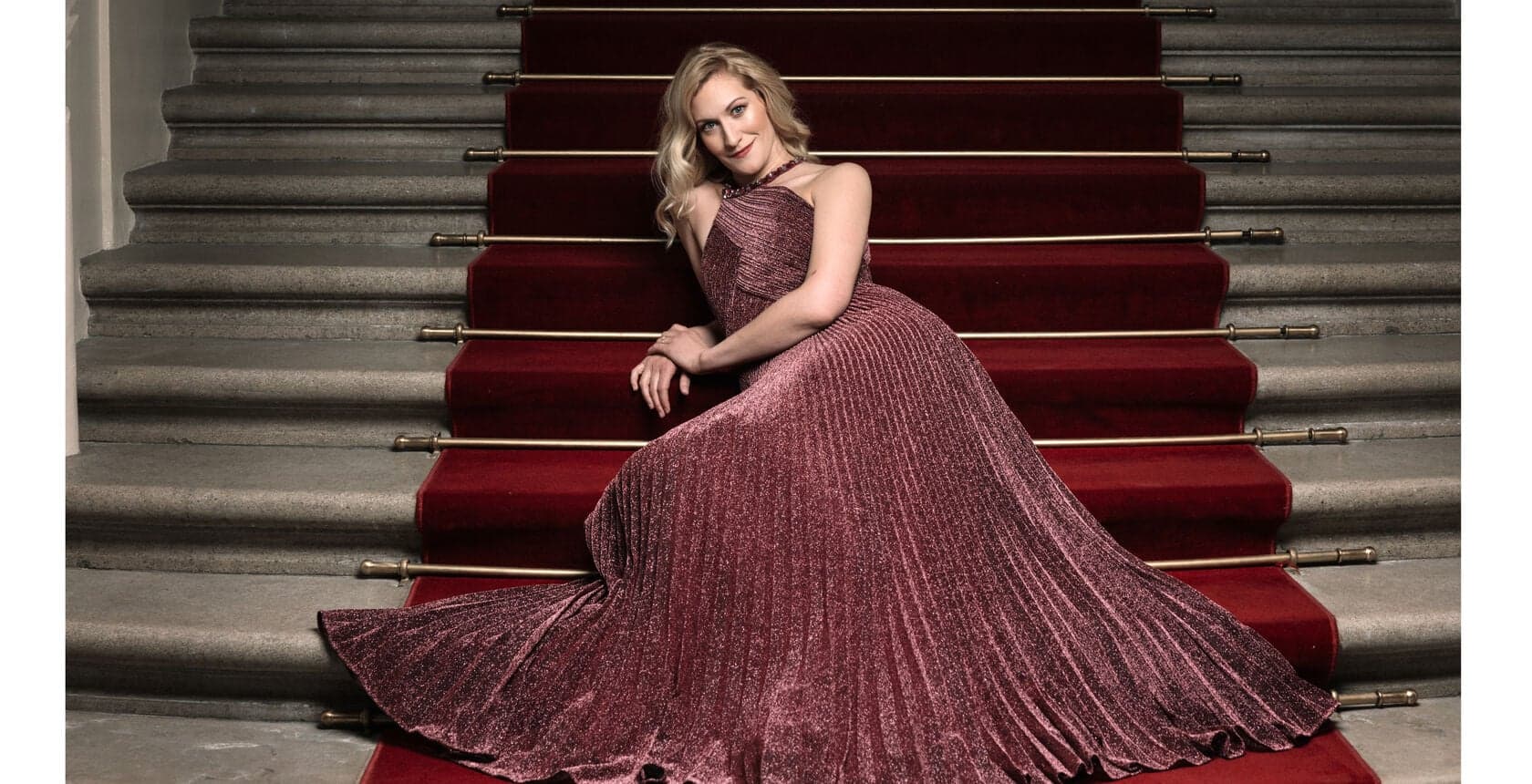 A woman in a long pink dress sits on red-carpeted stairs.