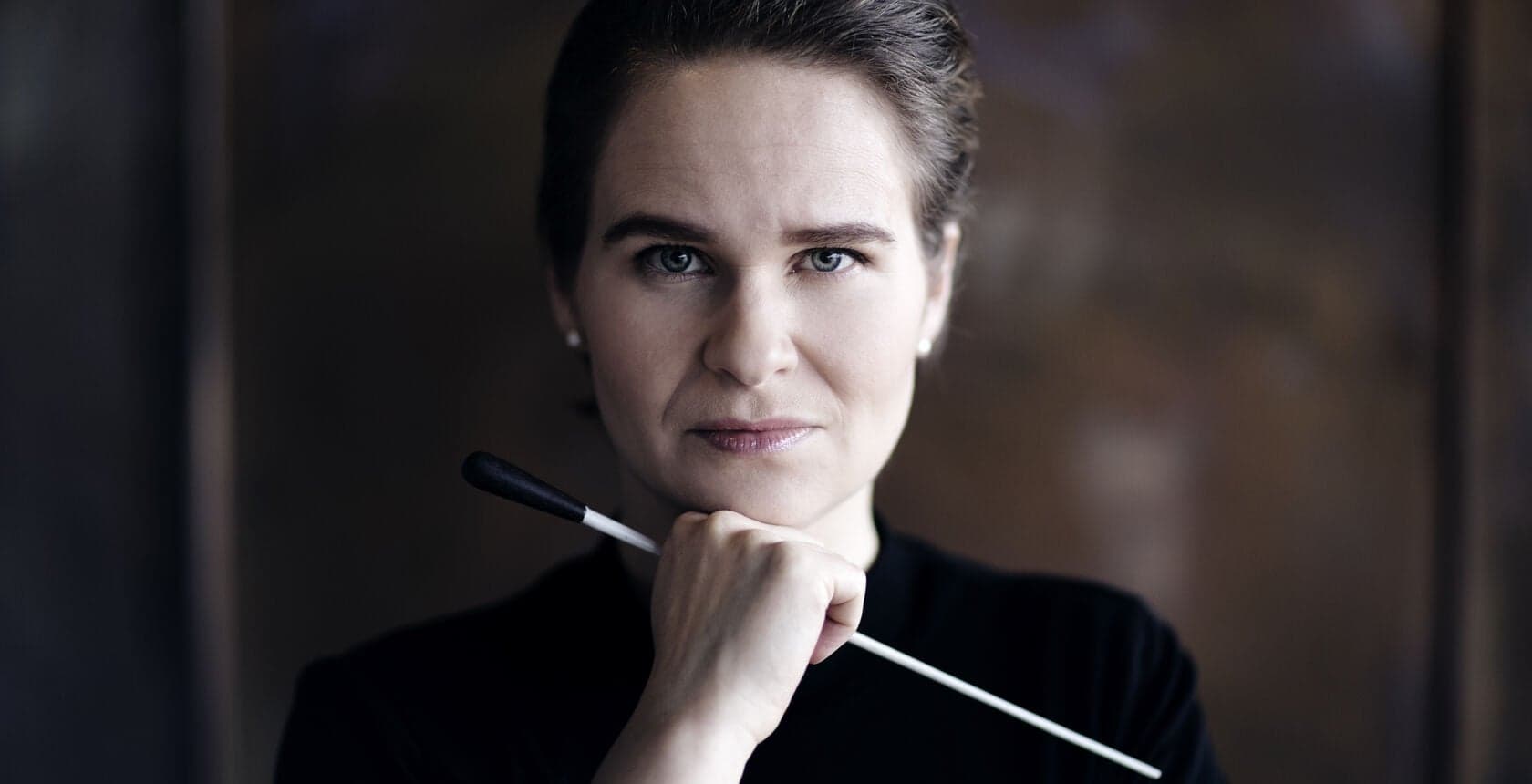 A woman holds a baton with a hand at her chin, against a dark background.