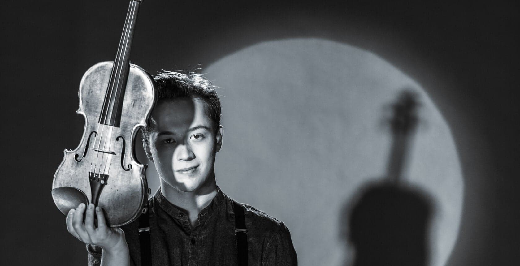 In black and white, a young man holds a violin with his shadow on a lit wall behind him.