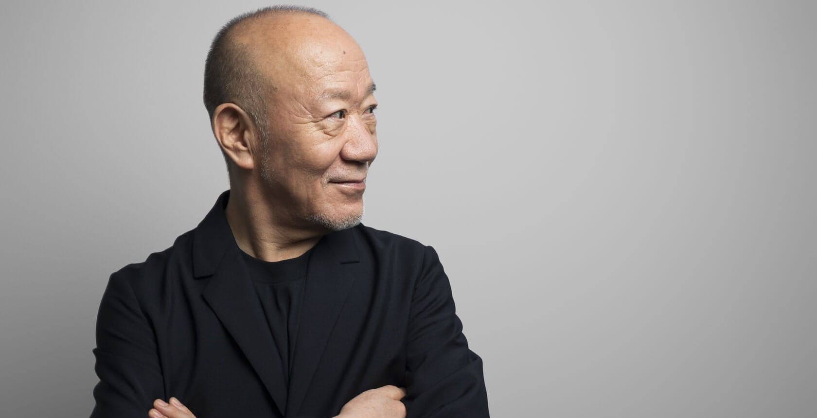A man in a dark jacket with his arms folded against a pale grey wall.