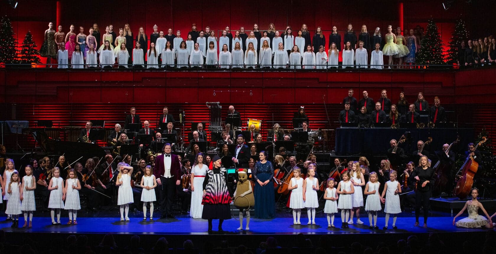 Choirs in rows on a stage with an orchestra, children in white at the front.