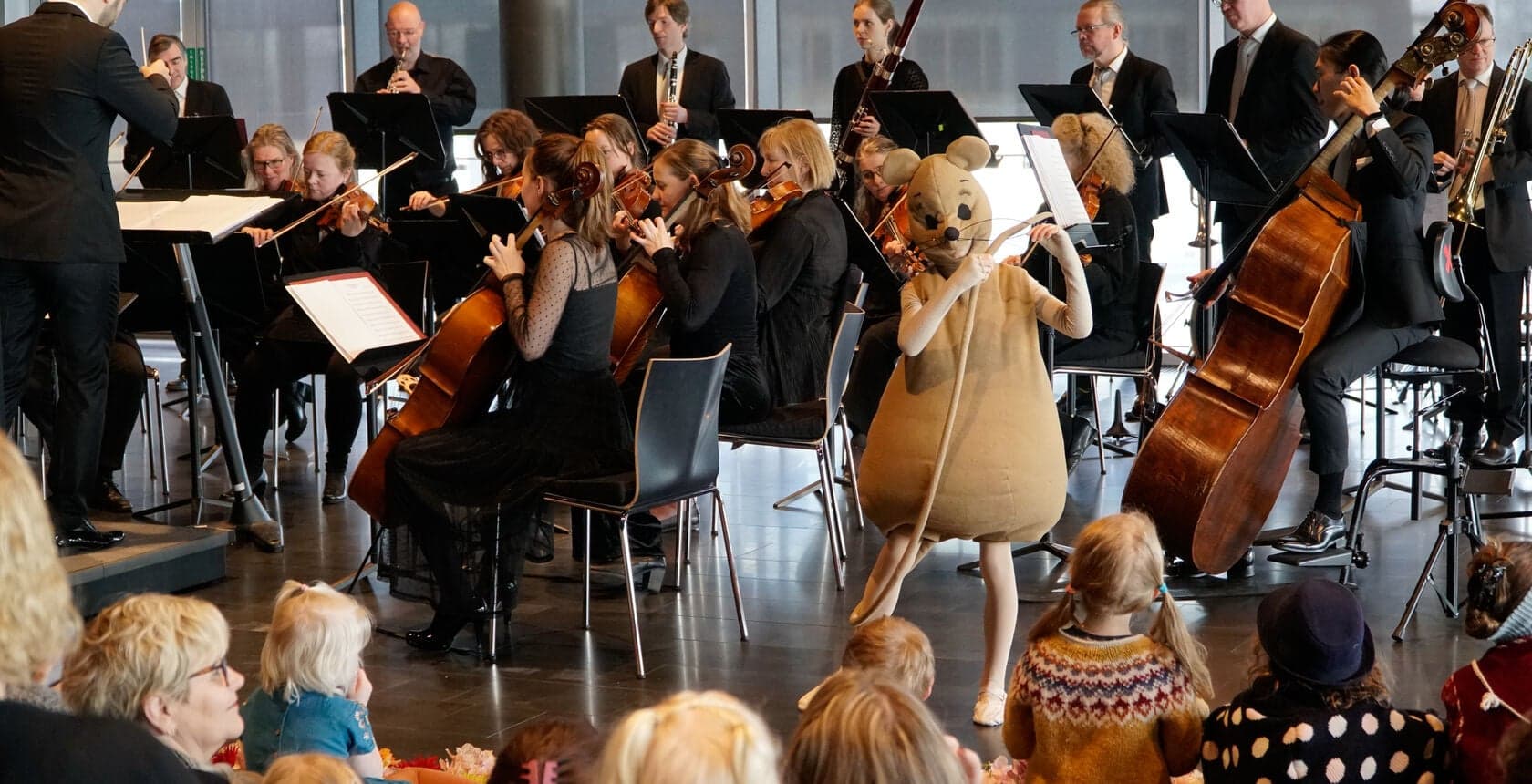 Musicians play to small children sitting on the floor, with a large soft-toy mouse among them.