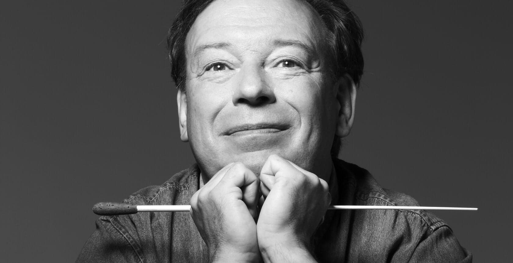 A black-and-white portrait of a smiling man resting his chin on his hands with a baton between them.
