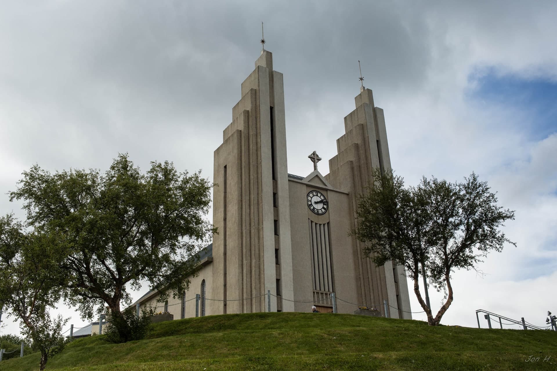 Akureyrarkirkja seen from the front up a grass slope, its two stepped towers standing either side of the clock with trees to left and right.