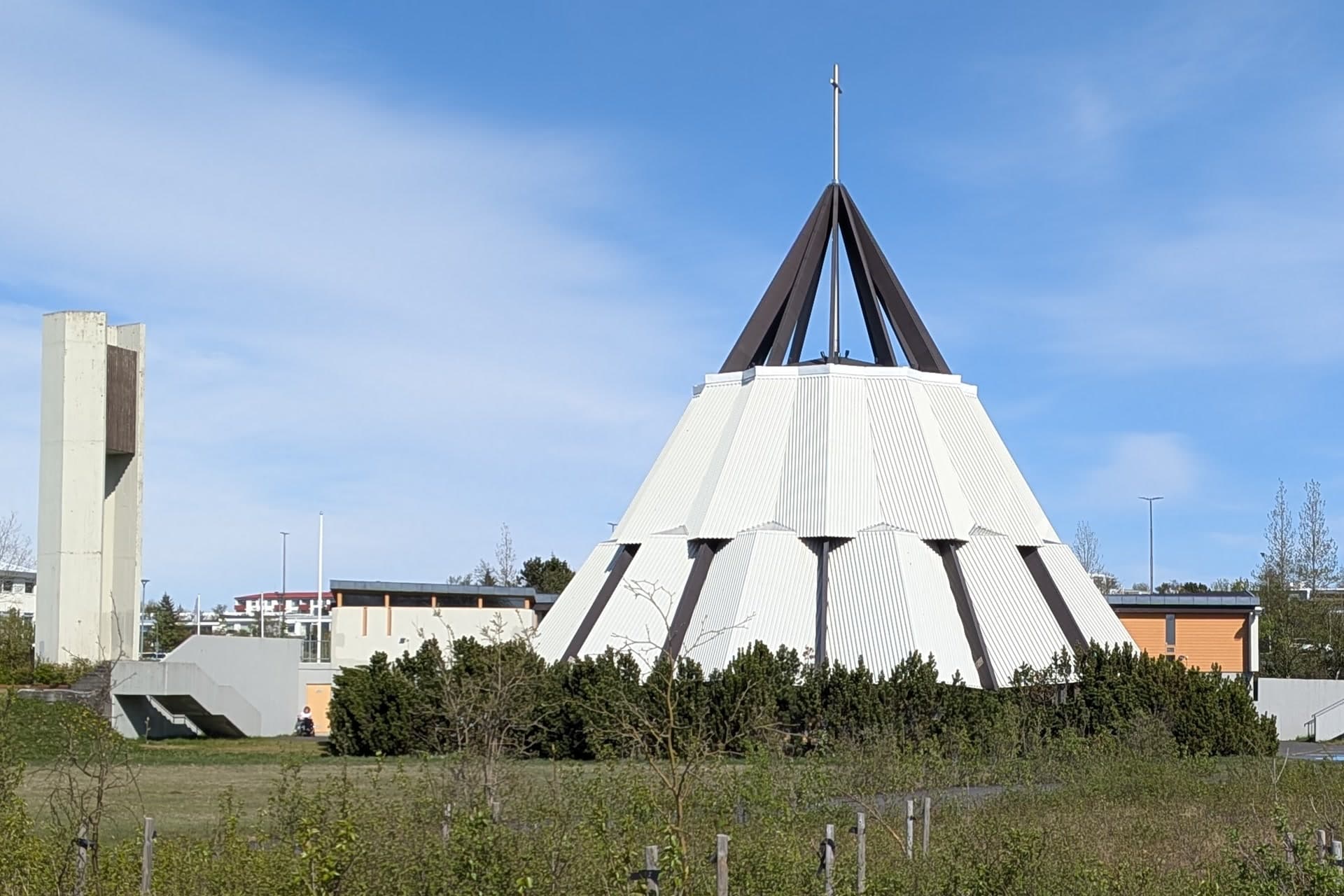 Breiðholtskirkja, hvít og keilulaga, þar sem dökkbrúnir bitar mætast í toppi undir krossi, og frístandandi klukknaturn úr steinsteypu til vinstri.