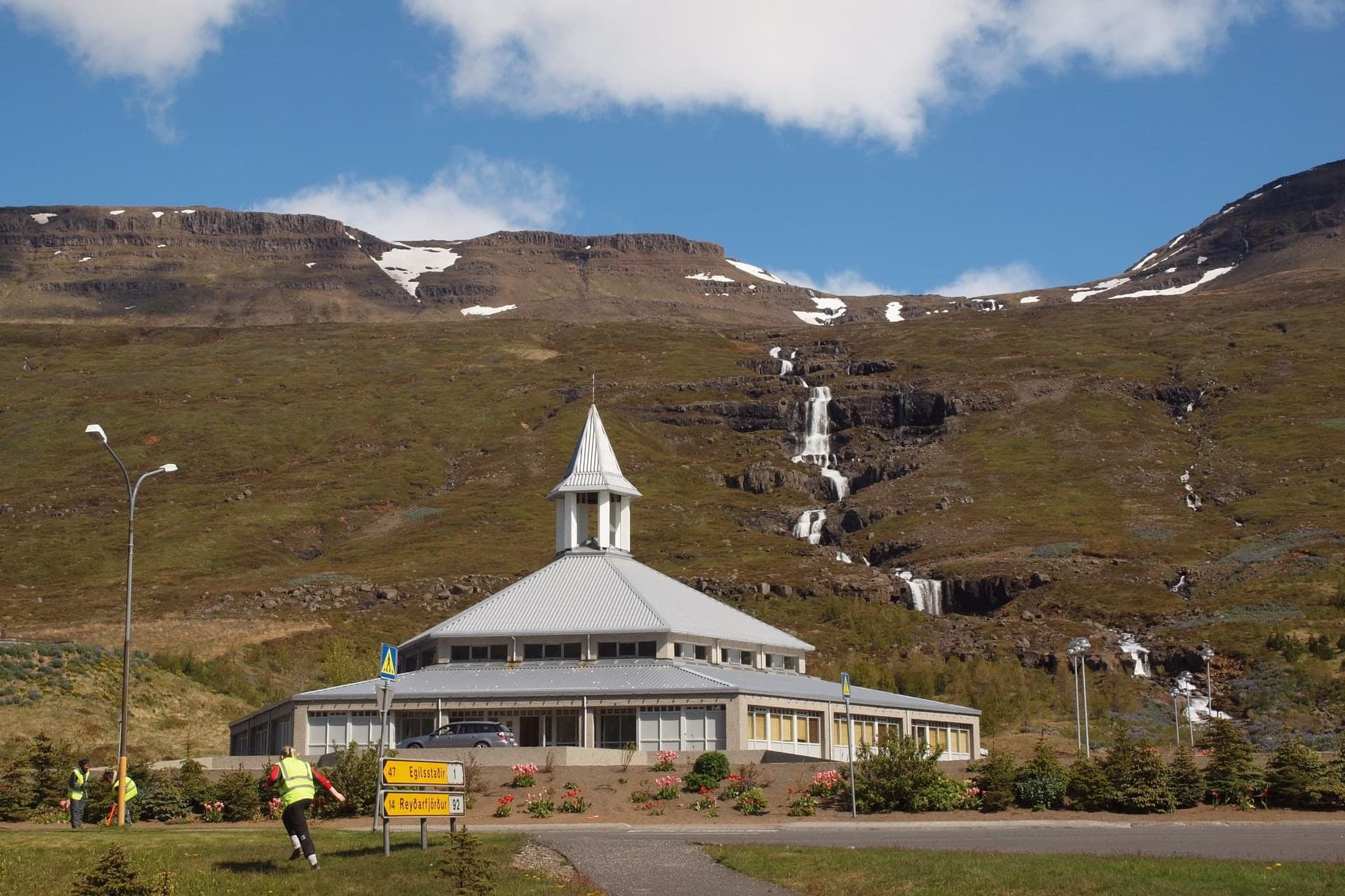 Eskifjarðarkirkja below a green mountainside with waterfalls, a low many-sided church with a pale metal roof and a white spire, with road signs and flower beds in front.