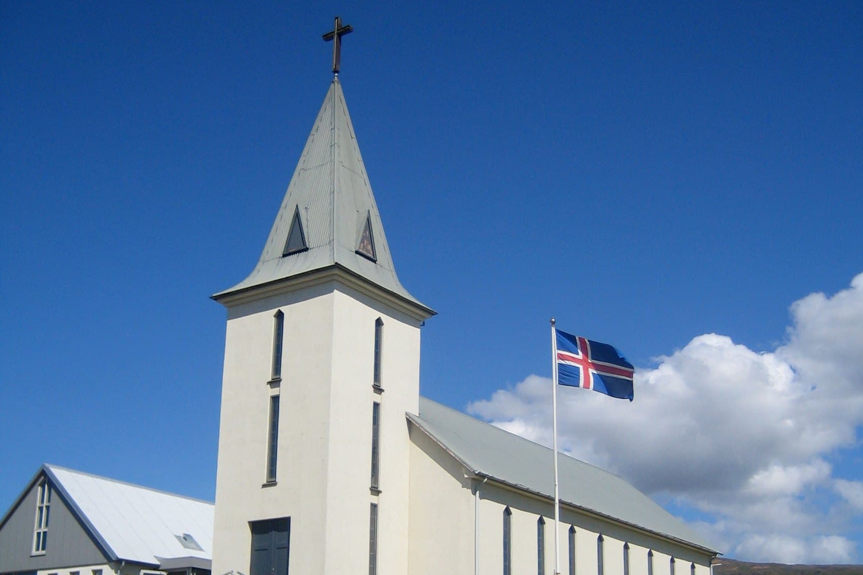 Hvammstangakirkja, hvítur turn með krossi á oddhvössu þaki, kirkjuskipið til hægri og íslenski fáninn við hún.