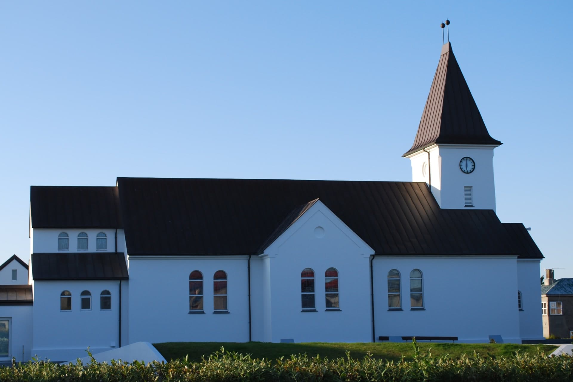 Keflavíkurkirkja frá hlið í sól, hvít kirkja með dökkbrúnu þaki og bogadregnum gluggum, og til hægri turn með klukku og oddhvössu þaki.