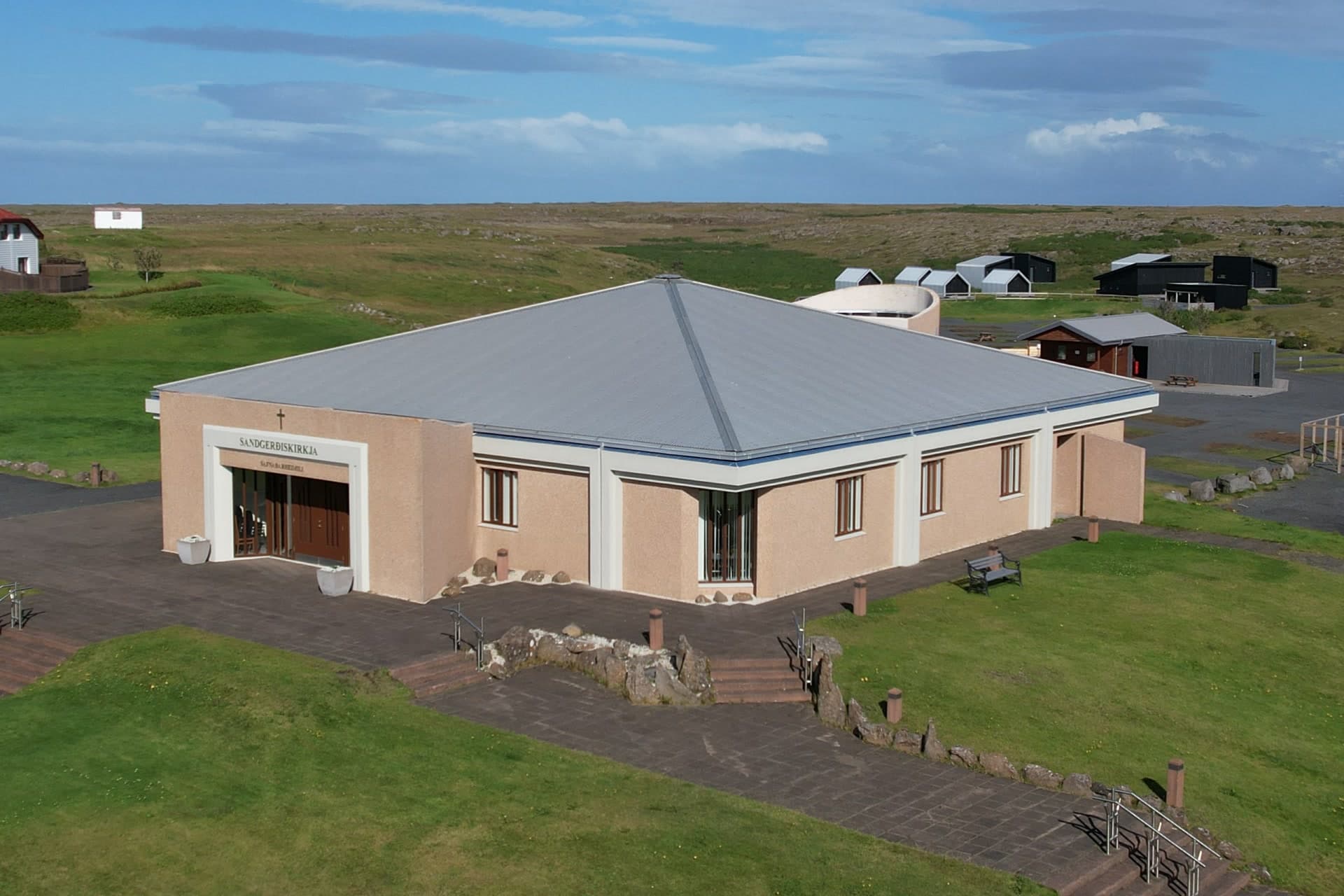 Sandgerðiskirkja from above, a square building with pale walls and a grey pyramid roof, the church's name over the entrance and steps and lawns in front.